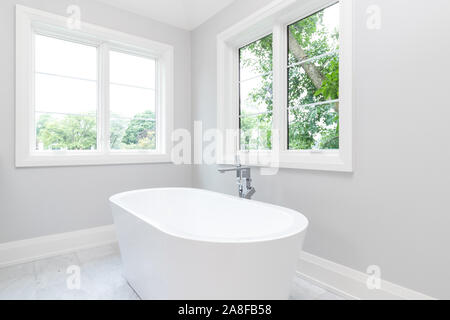 Eine schöne, weiße standalone Badewanne mit Chrom Hardware und schönen grünen Bäumen werden aus den Fenstern zu sehen. Stockfoto