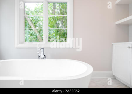 Detail geschossen von einem wunderschönen weißen standalone Badewanne mit Chrom Hardware und schönen grünen Bäumen werden aus den Fenstern zu sehen. Stockfoto