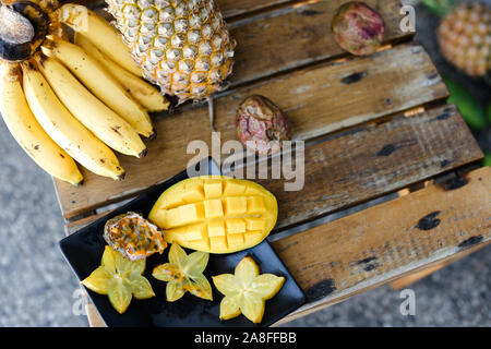 Karambolen mit Mango in schwarze Platte in der Nähe von Bananen und Ananas auf hölzernen Tisch. Stockfoto