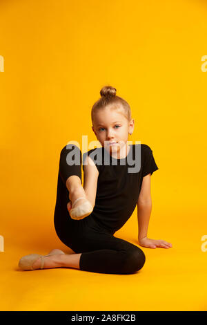 Volle Länge kleines Mädchen in schwarzen Body stretching Beine und auf der Suche nach weißen sitzen gegen hellen gelben Hintergrund während der Ausbildung Stockfoto