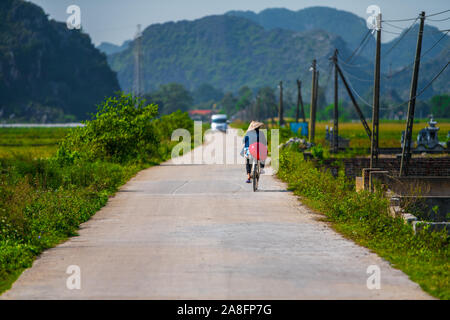 Ein lokaler Tam Coc resident Fahrten entlang einer kleinen Straße, neben Reisfeldern und wunderschöne Berglandschaft in Ninh Binh, Northern Vietnam - Herbst 2019 Stockfoto
