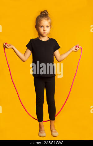 Volle Länge kleines Mädchen in schwarzen leotard Springen auf einem rosa Seil gegen hellen gelben Hintergrund. Stockfoto