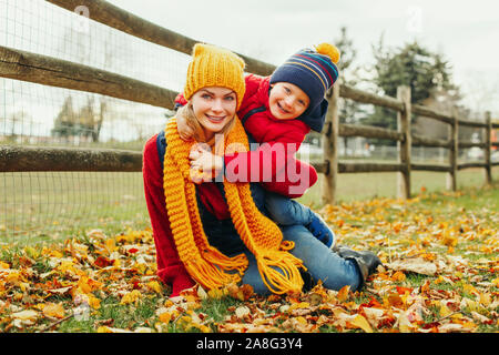 Kaukasische junge Mutter sitzt auf bodenanpassung cute adorable Toddler boy Sohn im Herbst park Outdoor mit gelb orange rot Blätter. Danksagung Stockfoto