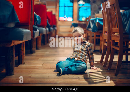 Cute adorable Junge drei Jahre alt Spaß im Cafe Restaurant. Kind sitzen auf dem Boden in der Öffentlichkeit. Freiheit der Meinungsäußerung und Verhalten für Stockfoto