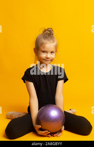 Gymnast Mädchen in Schwarz trico volle Höhe sitzt auf einer halben Seite mit einem Sport Ball auf gelbem Hintergrund Stockfoto