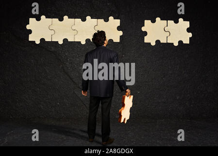 Ein Geschäftsmann Holding das fehlende Stück des Puzzles, ein unfertiges Puzzle hängt eine schwarze texturierte Wand Stockfoto