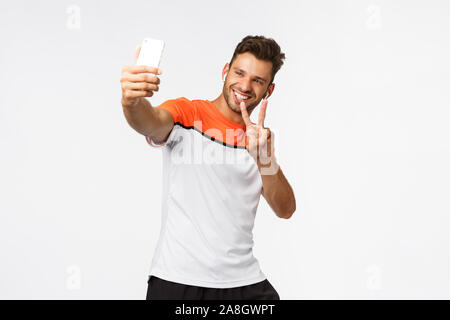LIfestyle, Sport und Menschen Konzept. Fröhliche stattlichen mascular männlichen Athleten in der Turnhalle, Verschleiß Sportswear, drahtlose Kopfhörer, wobei selfie auf dem Smartphone Stockfoto