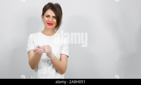 Schöne Kosmetikerin mit roten Lippenstift in Weiß medizinische Robe stellt Gummihandschuh an Hand gegen weiße Wand closeup kopieren Raum Stockfoto
