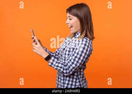 Profil von überrascht schöne Brünette Frau mit charmanten Lächeln in kariertem Hemd lesen Nachricht auf Handy, angenehm von guten Nachrichten überrascht. ind Stockfoto