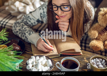 Mädchen in einem Pullover ist Schreiben eine Wunschliste, die in einem Notebook mit einem Becher Kaffee am Abend in der warmen Atmosphäre von Weihnachten. Gemütliche neue Jahr Stimmung Stockfoto