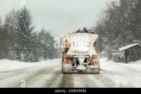 Schnee bedeckt orange Highway maintenance Streuer Fahrzeug auf rutschigem, starker Schneefall und Bäume im Hintergrund. Stockfoto