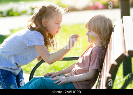 Ein kleines Mädchen ist die Anwendung aqua Make-up im Gesicht von einem 5 Jahre alten Jungen, der auf der Bank sitzt in einem Park. Stockfoto