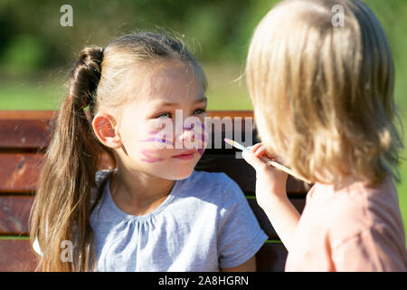 Ein kleiner Junge bringt aqua Make-up auf dem Gesicht eines 6-jährigen Mädchens, das auf der Bank im Park sitzt. Stockfoto