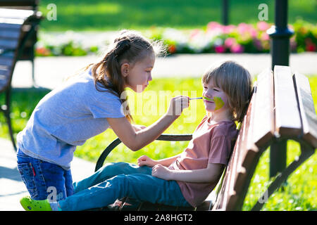 Ein kleines Mädchen ist die Anwendung aqua Make-up im Gesicht von einem 5 Jahre alten Jungen, sitzen auf einer Bank in einem Park. Stockfoto