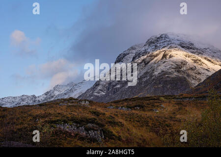 Dramatische Himmel über die schneebedeckten Gebirge durch die aufgehende Sonne und Herbstfarben im Tal unten. majestätischen Landschaft von Glencoe in den schottischen Highlands beleuchtet. Stockfoto