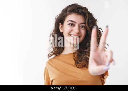 Lucky gefühlvolle charismatische optimistische junge Lockigen dunkelhaarigen Mädchen schwarze Haare Verlängerungshebel zeigt Ihnen Frieden Sieg Geste erreichen Erfolg top Stockfoto