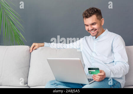 Online Shopping. Junger Mann studio Sitzen auf einem Sofa isoliert auf grau, Online Shopping auf Laptop per Kreditkarte zahlen für Erwerb Lächeln fröhlicher Stockfoto