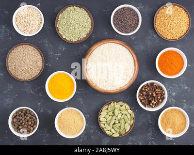 Reis, Piment, Pfeffer, Chili, Nelken, Bockshornklee, Kurkuma, Masala, Ingwer, Fenchel, Grüner Kardamom, Senfkörner in Schalen auf schwarzem Beton backgr Stockfoto