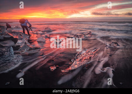 Fotograf bei Sonnenaufgang im Diamond Beach, Jokulsarlon, South Island. Bunte sunrise spiegelt sich auf dem nassen schwarzen Sand und das Eis. Stockfoto