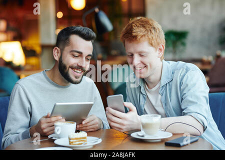 Zwei glückliche Kerle mit mobilen Gadgets beobachten neugierig Material in Smartphone Stockfoto