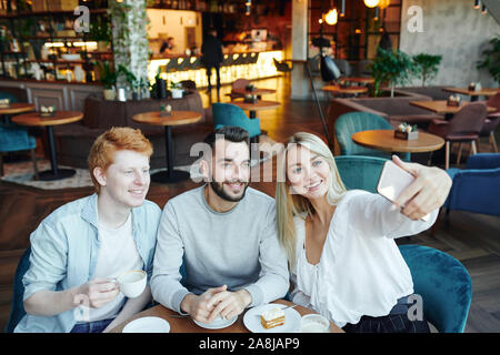Drei glückliche junge Freunde sitzen durch Tisch im gemütlichen Cafe und selfie Stockfoto