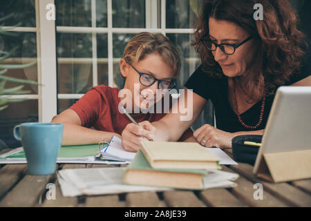 Junge Schüler Hausaufgaben zu Hause mit Schule Bücher, Zeitung und digitale Pad von seiner Mutter geholfen. Mom schreiben auf dem copybook Lehre seinen Sohn.