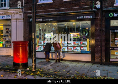 Zwei Frauen auf der Suche nach Details in einem beleuchteten Fenster der Immobilienmakler in der Nacht, auf Kings Lynn Dienstag Markt. Stockfoto