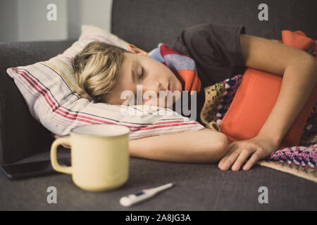 Kranker Junge Schlafen mit Wolldecke und Wärmflasche. Traurig jugendlich mit der Grippe liegt allein in einem kalten Wintertag. Kind mit saisonalen infizieren. Stockfoto