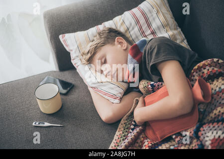 Kranker Junge Schlafen mit Wolldecke und Wärmflasche. Traurig jugendlich mit der Grippe liegt allein in einem kalten Wintertag. Kind mit saisonalen infizieren. Stockfoto