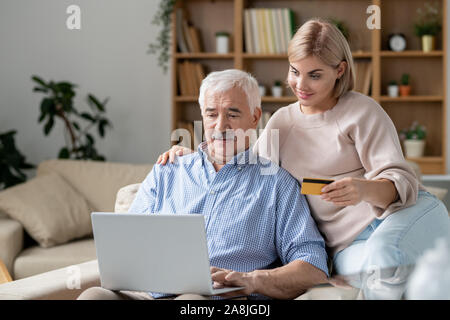 Hübsche junge Frau mit Kreditkarte sitzend durch ihr älterer Vater online einkaufen Stockfoto