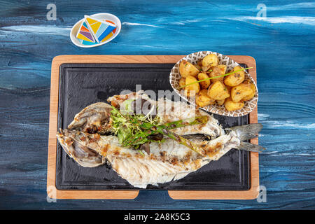 Den gegrillten Wolfsbarsch auf dem Holzbrett. Den gegrillten Wolfsbarsch, serviert auf einem Teller mit verschiedenen Saucen und mit Petersilie und Zitrone eingerichtet. Stockfoto