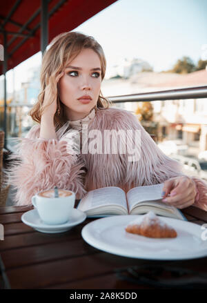 Süße Mädchen im Restaurant sitzen, Kaffee trinken (Cappuccino) und lesen ein Buch. Portrait von stilvollen junge Frau im Cafe, das Tragen von Kleidung und Pelzmantel Stockfoto