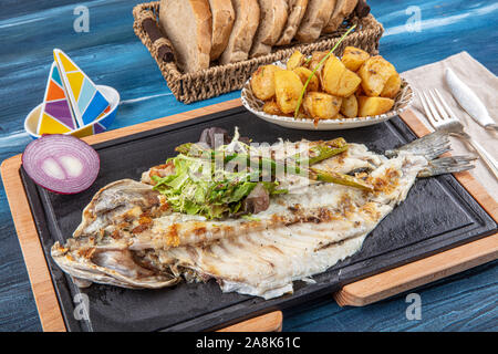 Den gegrillten Wolfsbarsch auf dem Holzbrett. Den gegrillten Wolfsbarsch, serviert auf einem Teller mit verschiedenen Saucen und mit Petersilie und Zitrone eingerichtet. Stockfoto