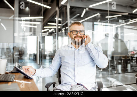 Senior Manager zuversichtlich, am Telefon zu sprechen, während am Arbeitsplatz sitzen im modernen Büro Stockfoto