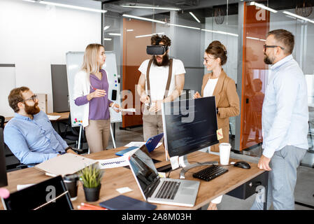 Gruppe von verschiedenen Kollegen während kleine Konferenz im Büro, kreativ bärtiger Mann, der versucht, ein neues Produkt mit Virtual reality Brillen Stockfoto