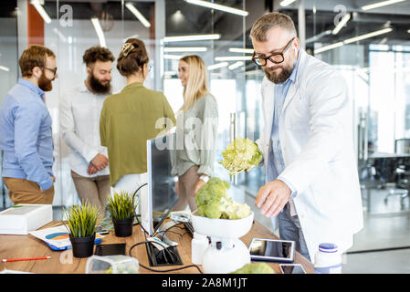 Leitende Ernährungsberaterin arbeiten auf einer Diät Plan am Arbeitsplatz mit Computer und frische Lebensmittel im Büro mit jungen Kolleginnen und Kollegen im Hintergrund Stockfoto
