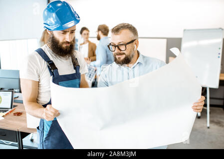 Oberpolier und Arbeiter in Latzhosen, die am Projekt mit Blueprints im Büro Stockfoto