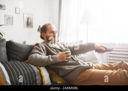 Seitenansicht Portrait von mittlerem Alter bärtiger Mann vor dem Fernseher zu Hause und Schaltkanäle über Fernbedienung und entspannen Sie auf bequemen Couch Stockfoto