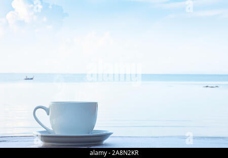 Heißer Kaffee in eine weiße Tasse mit Untertasse auf einem Holztisch Hintergrund verschwommen Boot und das Meer. Stockfoto