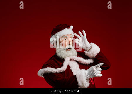 Taille bis Portrait von Santa Claus überrascht, die auf der Seite und Anpassung der Brille, während gegen den roten Hintergrund posiert im Studio, kopieren Raum Stockfoto