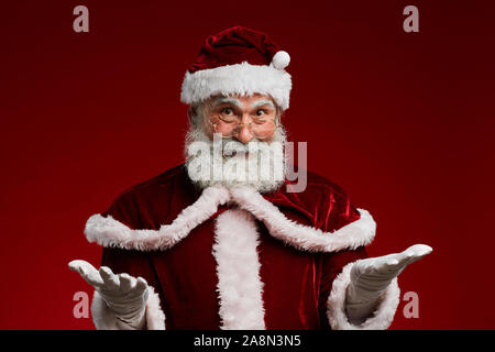 Taille bis Portrait Of Smiling Santa Claus begrüßt die Gäste halten die Arme im Stehen gegen die roten Hintergrund im Studio, Kopie Raum Stockfoto