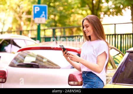 Schuss der jungen Frau, die auf der Straße neben ihrem Auto und Zahlen für Parken per Handy. Stockfoto