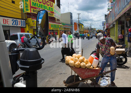 HIGUEY, Punta Cana, Dominikanische Republik - 25. JUNI 2019: Mann mit Schubkarre verkaufen frische Kokosnüsse. Blick auf typische Südamerikanische Stadtbild - Straße mit Autos, Stockfoto