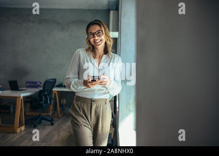 Fröhliche Frau, die mit ihrem Telefon. Mit Handy in der Hand Geschäftsfrau in die Kamera schauen und lächeln. Stockfoto