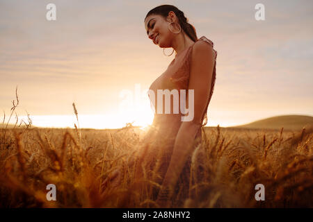 Junge Frau im Weizenfeld mit Sonnenuntergang im Hintergrund. Frauen in einem landwirtschaftlichen Gebiet während des Sonnenuntergangs. Stockfoto