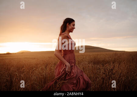 Junge Frau wandern im Weizenfeld ihr Kleid Holding. Frau in der Landschaft Weizenfeld bei Sonnenuntergang. Stockfoto