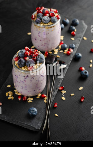 Gesundes Frühstück Konzept. Gläser mit hausgemachtem Joghurt mit Blaubeeren, Granatapfel Samen und Hafer auf schwarzem Hintergrund. Vertikale. Stockfoto