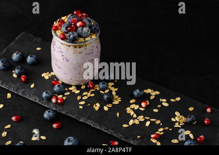 Gesunde fitness food Konzept. Glas hausgemachten Joghurt mit Blaubeeren, Granatapfel Samen und Hafer auf schwarzem Hintergrund. Vorderansicht, Kopie Raum Stockfoto