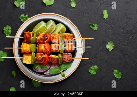 Paneer Tikka auf Spieße in schwarz Schüssel am dunklen Schiefer Hintergrund. Paneer Tikka ist eine indische Küche Teller mit gegrillten Paneer mit Gemüse und Käse Stockfoto