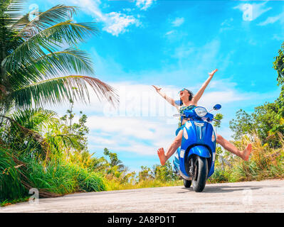 Crazy lustige Frau mit fliegenden Haare Motorrad fahren auf einem blauen Himmel und grüne Tropen Hintergrund. Junge bizarre Mädchen mit dunklen Haaren in Sonnenbrille auf ein Stockfoto